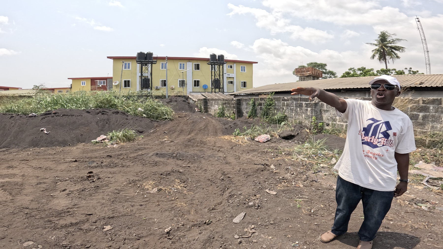 A man standing by a stone wall and indicating the dark ground of the yard